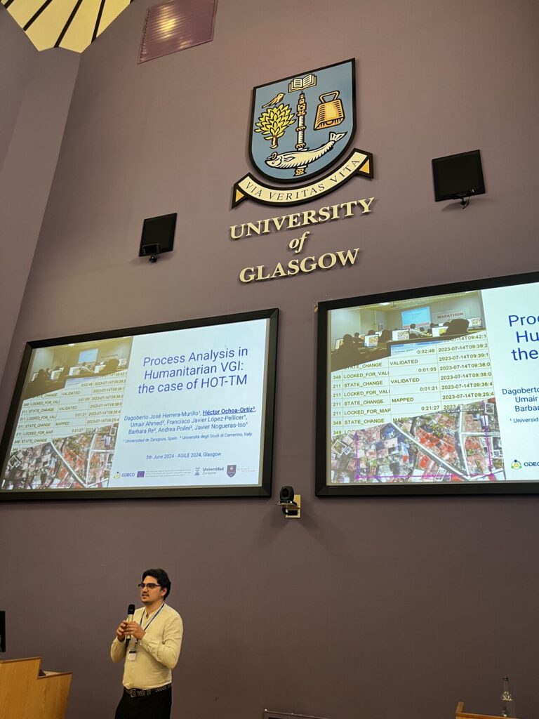 A big wall with two screens showing a presentation, on the bottom of the picture, a man is presenting with a microphone