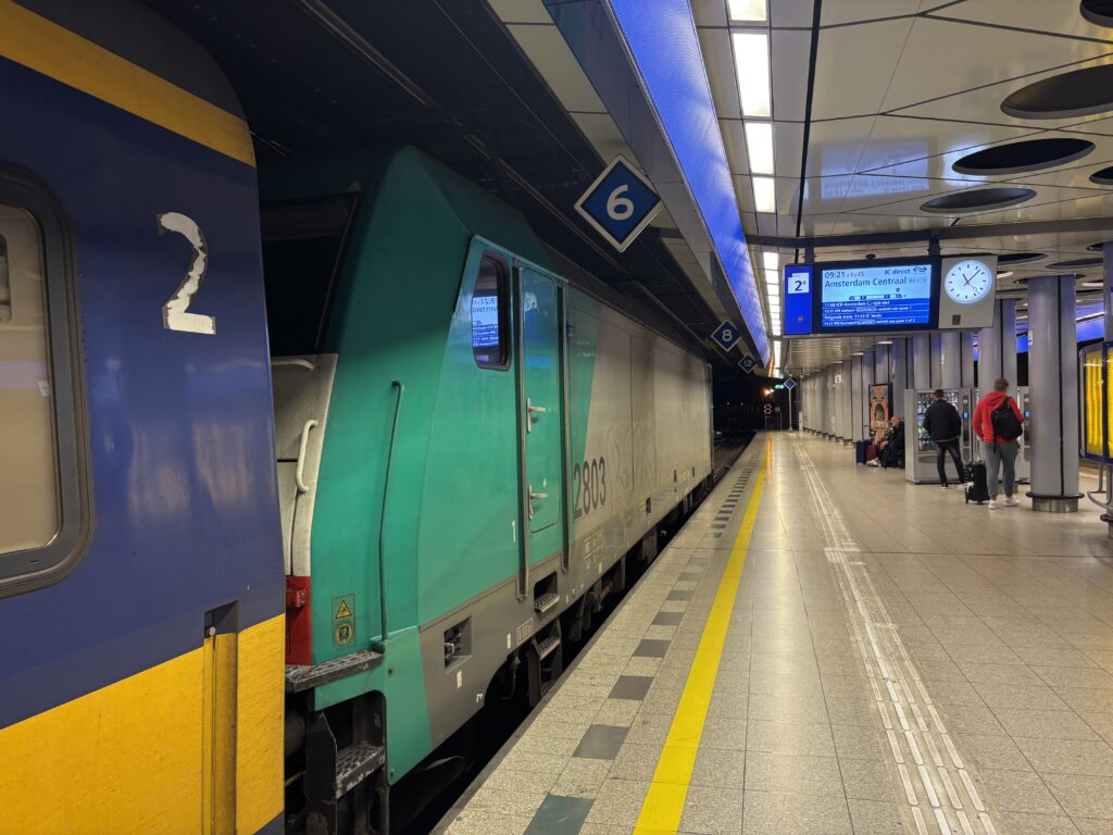 A locomotive followed by some train carriages, as seen from the platform edge. The screens shows a delay of 1 hour and 45 minutes of the current train.