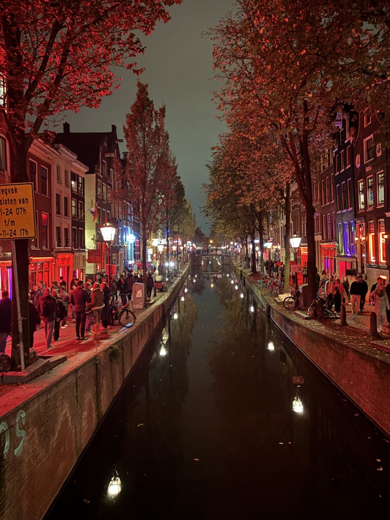 A canal at night surrounded by trees, and people packed streets with brick buildings that have red lighted windows