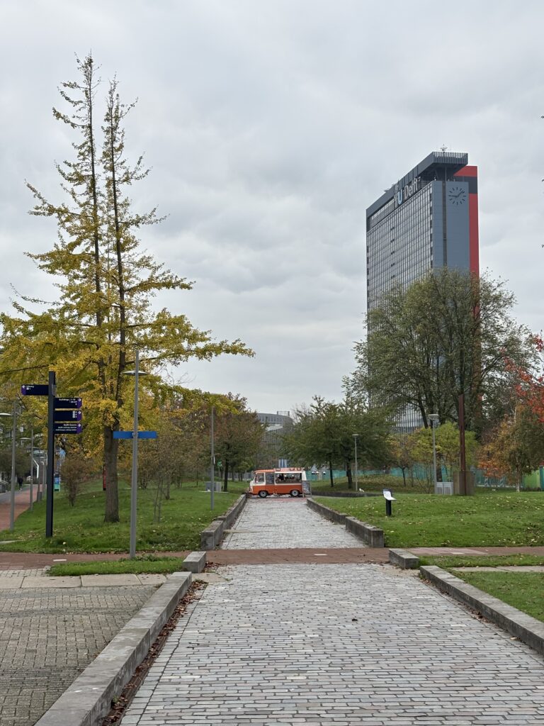 A long shot, with a food truck and a tall building in the background. It is all surrounded by greenery.