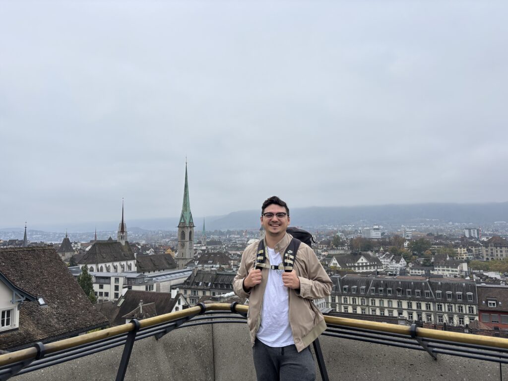 A man in the foreground, posing. On the background, a city landscape. The man is on an elevated position, where a big part of the city can be seen.