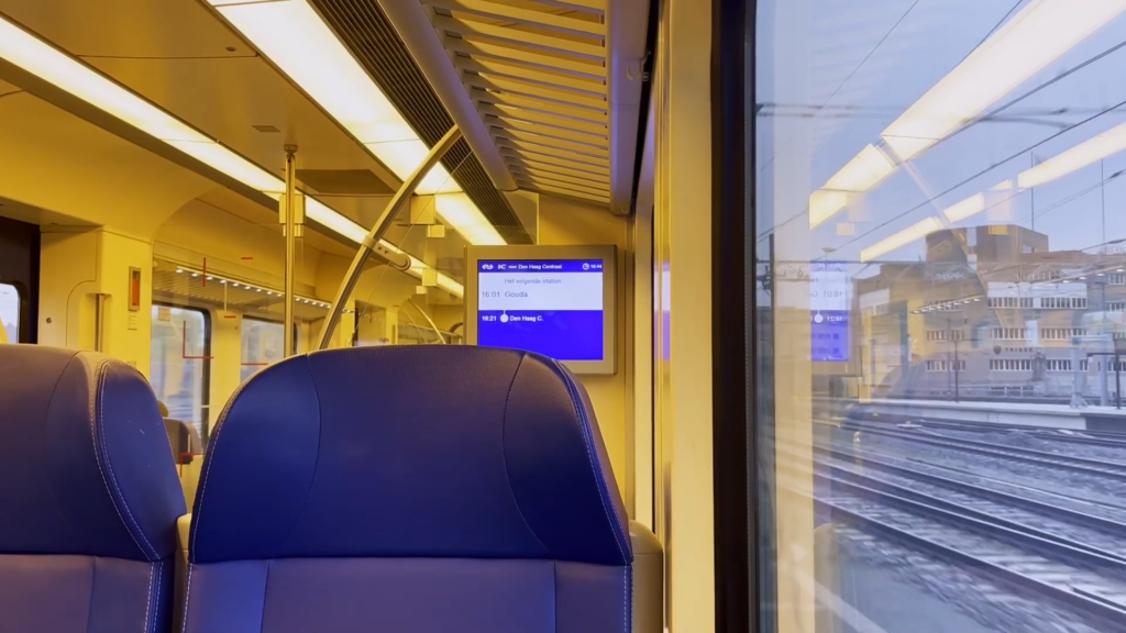 Interior view of a train, with padded seats, a screen displaying the next stops, and a window view showing multiple tracks